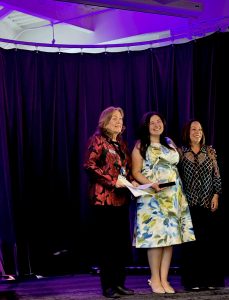 Photo of woman in a dress with an award flanked by two other women on a stage.