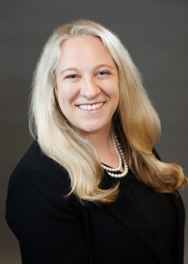 Headshot of a woman in a black blazer wearing pearls with shoulder length blond hair.