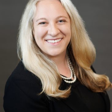 Headshot of a woman in a black blazer wearing pearls with shoulder length blond hair.