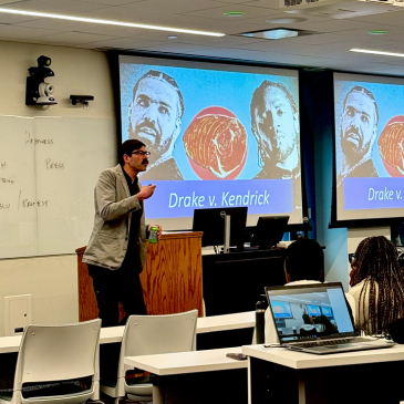 Professor Anthony Moffa stands in the front of the classroom before a powerpoint slide that says Drake vs. Kendrick