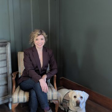 Photo of woman seated in chair with service dog lab lying next to her. Both are looking at the camera.