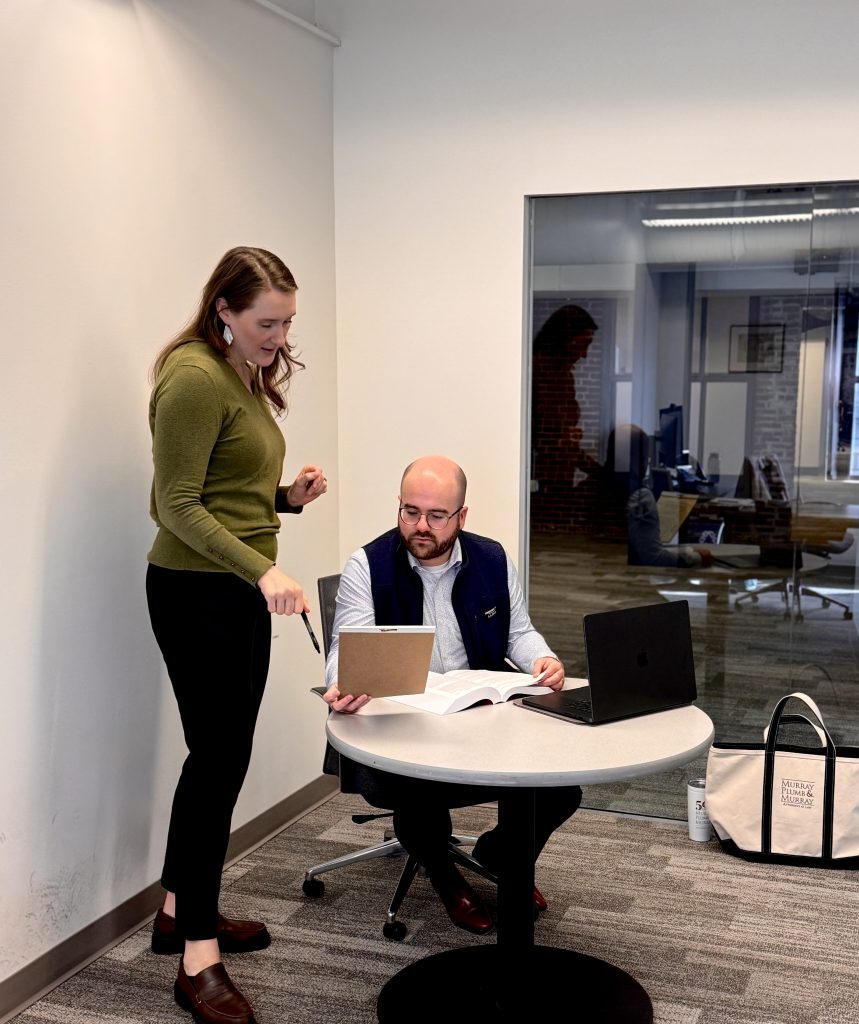 A woman stands over a seated man and they are both looking at a legal pad.