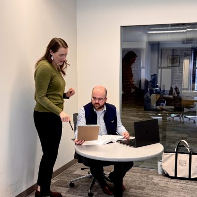 A woman stands over a seated man and they are both looking at a legal pad.