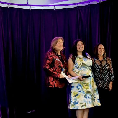 Photo of woman in a dress with an award flanked by two other women on a stage.