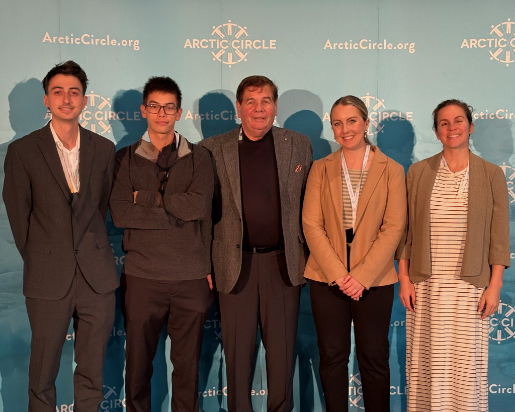 Photo of four students standing with Professor Norchi in front of a sign that says "Arctic Circle."