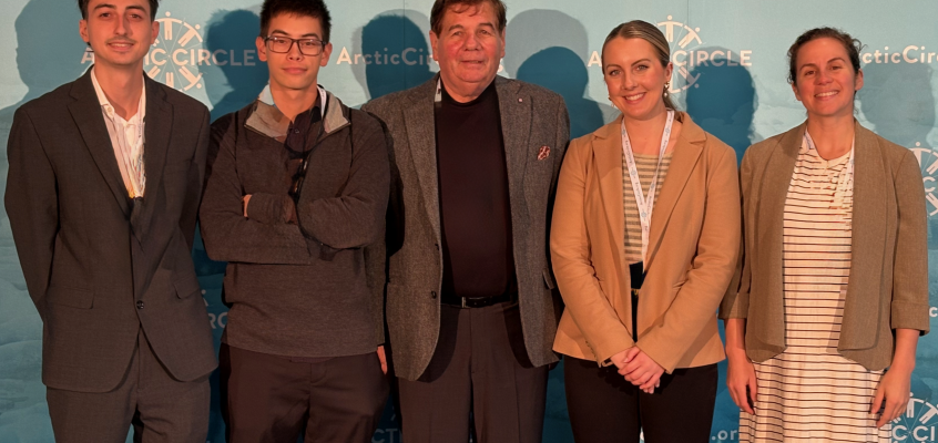 Photo of four students standing with Professor Norchi in front of a sign that says "Arctic Circle."