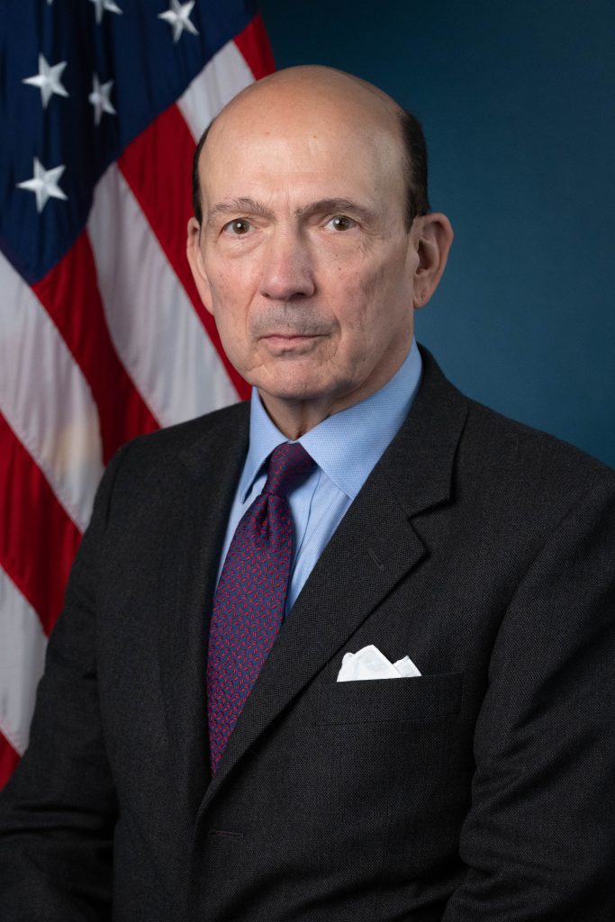 Photo of man in a suite in front of a U.S. flag.